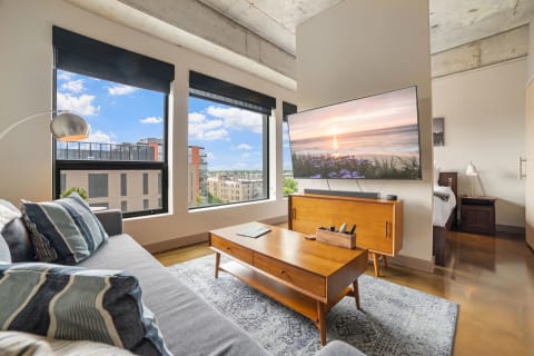 Contemporary living room with a gray sofa, wooden coffee table, and large windows showcasing a city view.