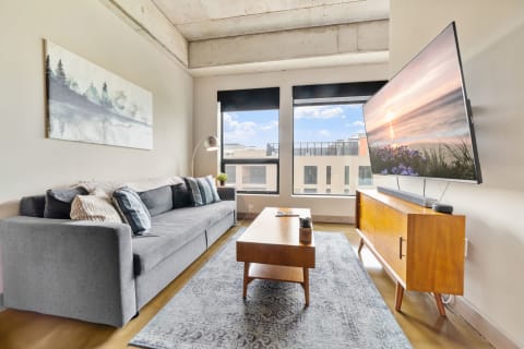 Modern living room with a grey sectional sofa, large TV, and sunlight streaming in through windows.