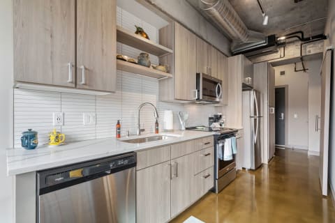 A modern kitchen featuring wooden cabinets, stainless steel appliances, and decorative pottery on open shelves.