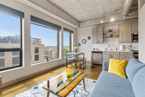 A contemporary living room with large windows, a blue sofa, a wooden coffee table, and a dining area.