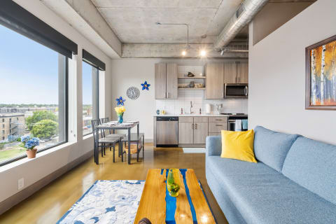 Interior of a contemporary apartment showcasing a blue couch, a dining table, and kitchen area with large windows.