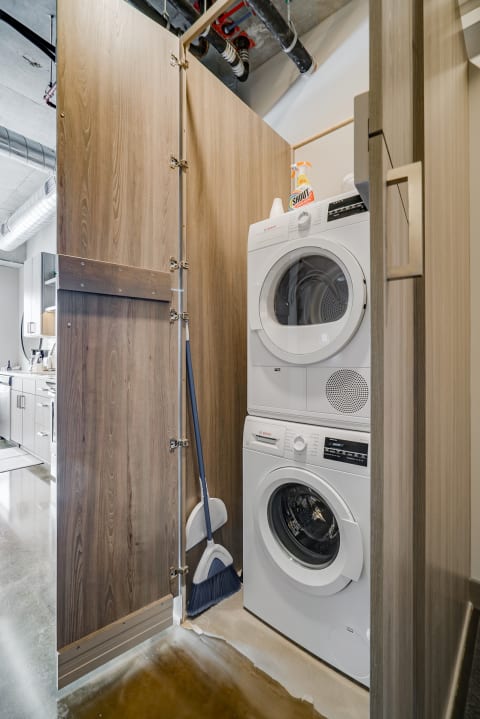 A compact laundry area with a stacked washer and dryer, wooden partition, and cleaning supplies.