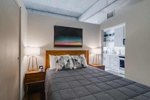 A bedroom featuring a grey quilted bed, geometric pillows, wooden nightstands with lamps, and a small kitchen area visible in the background.