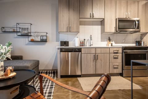 Interior view of a stylish kitchen with light wood cabinets and stainless steel appliances, connected to a cozy living area.