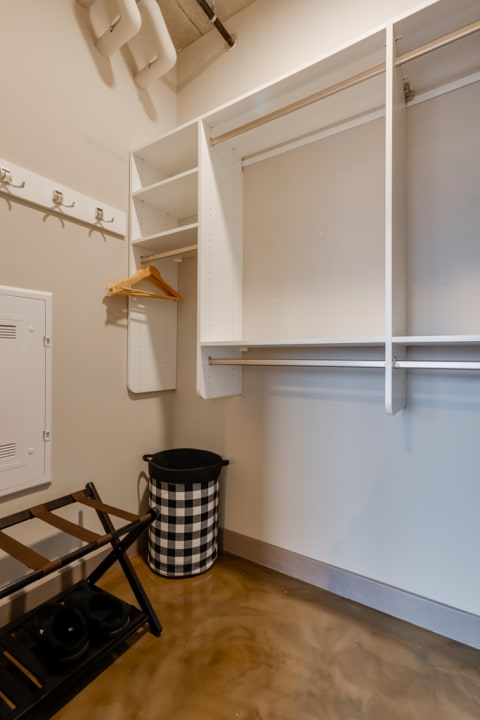 A minimalist closet space with white shelving, clothing hooks, a black and white checkered laundry basket, and a folding rack on a polished floor.