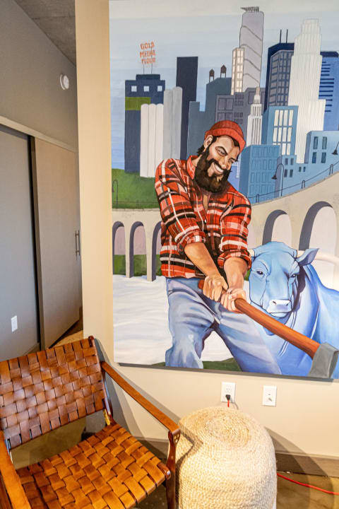 A colorful mural showing a muscular man in a red plaid shirt swinging a tool next to a blue cow and a city skyline.