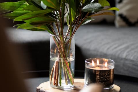 A glass vase with green leaves and a lit candle on a wooden slice near a textured gray couch.