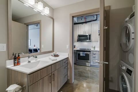 A modern bathroom featuring a white countertop, mirror, and an adjoining kitchen with laundry appliances.