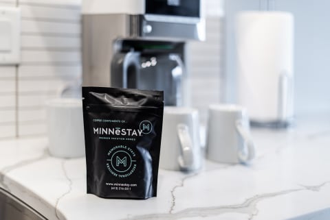 A black coffee bag labeled MINNēSTAY on a marble kitchen countertop with white mugs in the background.