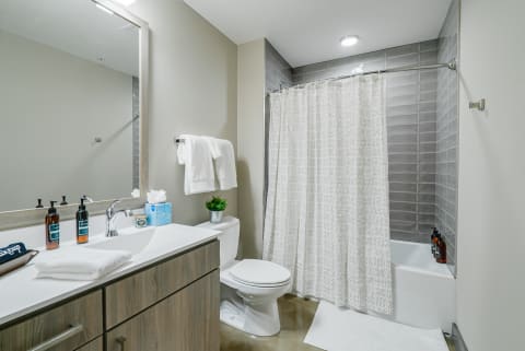 Modern bathroom with a wood vanity, white countertop, and gray tiled shower area.