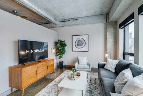 A stylish living room featuring a concrete ceiling, modern furniture, and a large TV with 'MINNESTAY' logo.