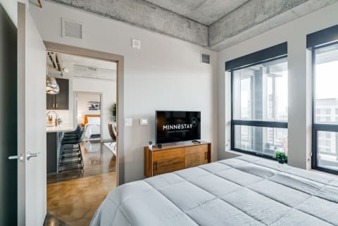A modern bedroom featuring a bed, TV, and large windows with a view of a kitchen area.