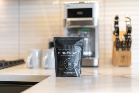 A black coffee bag from Minnestay on a kitchen countertop with a coffee maker and mugs in view.