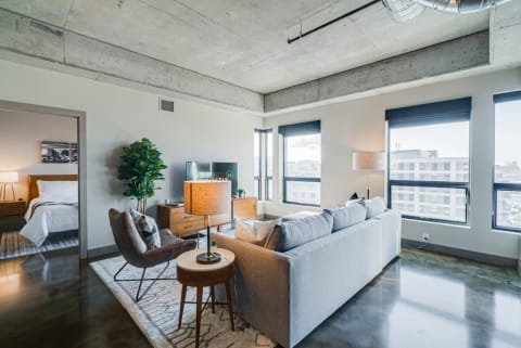 Modern living space with light gray sofa, warm lighting, and large windows showing a city view.