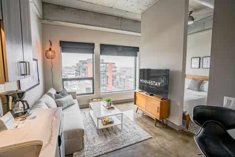 A modern studio apartment featuring a gray sectional sofa, a coffee table, and large windows revealing a city view.
