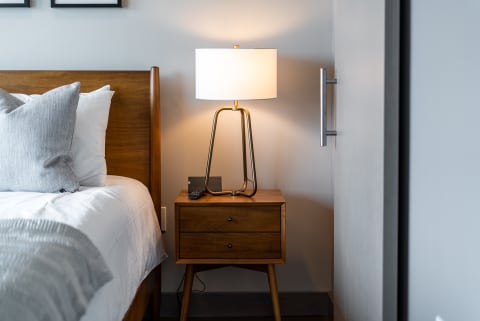 Warm bedroom corner with a wooden bed frame, decorative pillows, and a modern nightstand with a lamp.