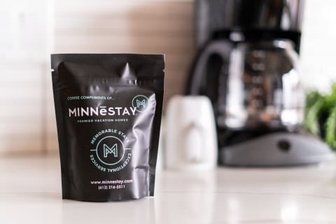 A bag of coffee from MINNESTAY displayed in a kitchen with a coffee maker and a white mug in the background.