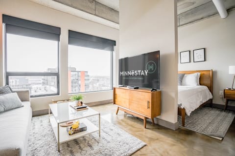 A modern studio apartment featuring a living space, a bed area, and large windows with city views.