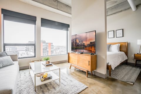 A modern loft-style living room with a sofa, coffee table, television, and a bedroom view.