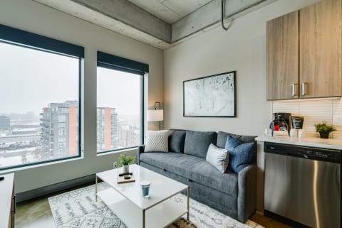 Modern living room with gray sofa, large windows, and a kitchen area.