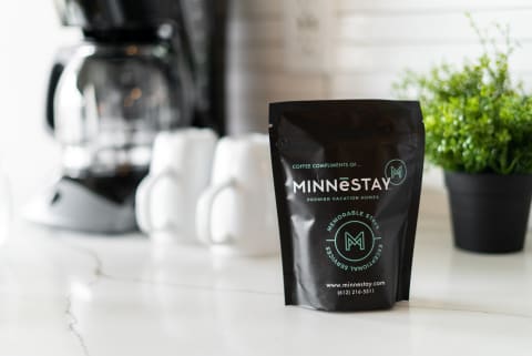 A black coffee bag from MINNESTAY next to a coffee maker and white mugs on a marble countertop.