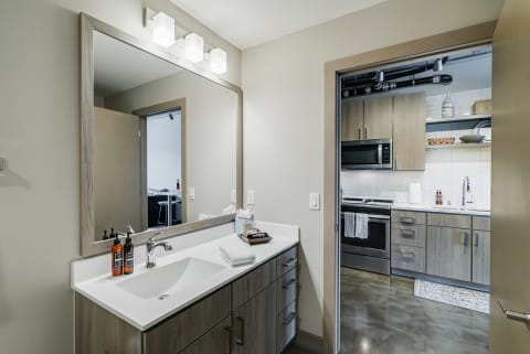 Interior view of a modern bathroom with a mirror and light fixtures, leading to a kitchen.