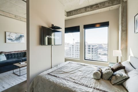 Modern bedroom and living area with large windows and city view, featuring a bed, TV, and sofa.