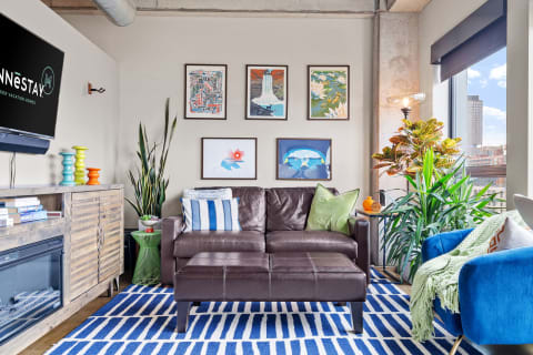 Modern living room featuring a brown leather couch, colorful cushions, vibrant artworks, and green plants.