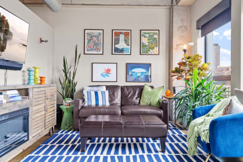 A contemporary living room featuring a brown leather sofa, colorful artwork on the walls, and a bright blue and white striped rug.