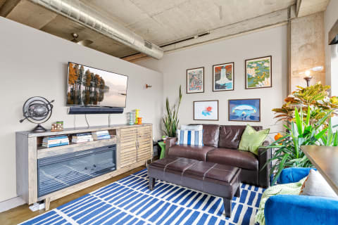 A modern living room featuring a brown leather couch, wooden media console with a TV, colorful art on the walls, and decorative plants.