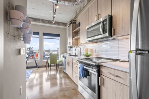 A contemporary kitchen with light wood cabinetry and stainless steel appliances, featuring large windows overlooking the city.