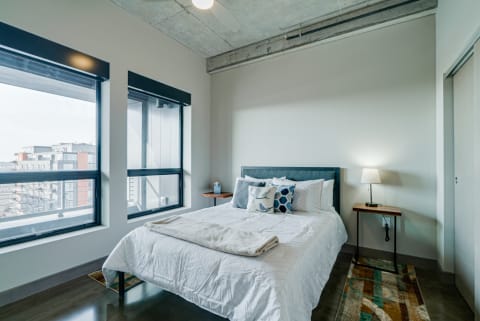 Modern bedroom with a bed, decorative pillows, and large windows showing an urban landscape.