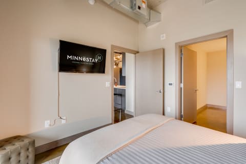 Stylish bedroom with a flat-screen TV showcasing MINNēSTAY logo, clean decor, and light-colored bedding.