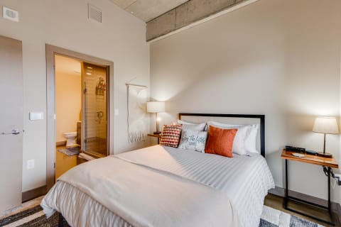 Cozy modern bedroom with white linens, decorative pillows, and a view of the bathroom.