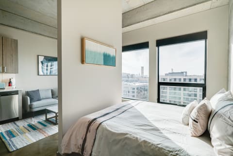 Interior of a stylish studio apartment featuring a bed, kitchenette, and large windows with city views.
