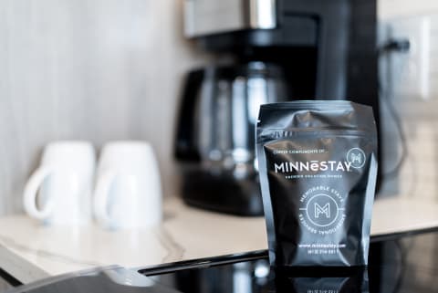 A bag of MINNéSTAY coffee placed on a kitchen countertop with white mugs in the background and a coffee maker.