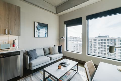 A contemporary living room with a gray sofa, coffee table, and large windows showing city views.