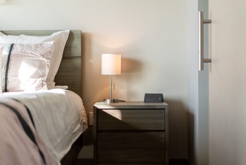 A cozy bedroom featuring a made bed, a lamp on a nightstand, and a modern electronic device.