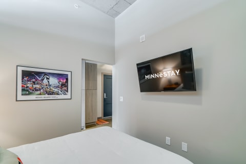 Interior view of a stylish hotel room with a bed, wall art, and a TV displaying 'MINNESTAY.'