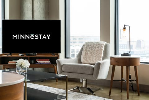 A modern living room with an armchair, side table, lamp, and a TV displaying the MINNESTAY logo.