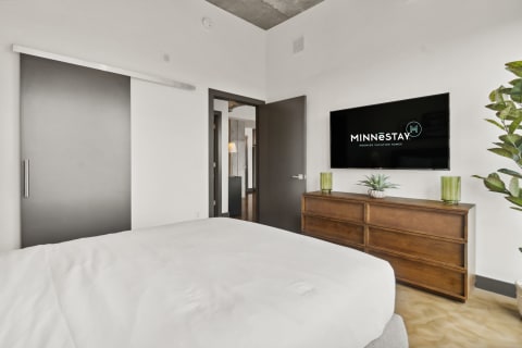 A modern bedroom featuring a white bed, wooden dresser, and mounted TV with MINNESTAY logo.