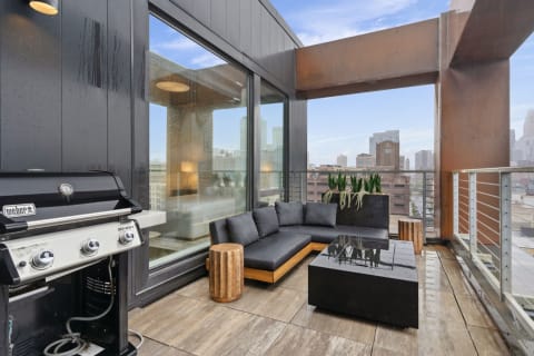 Modern balcony with sectional sofa, coffee table, and grill overlooking a cityscape.
