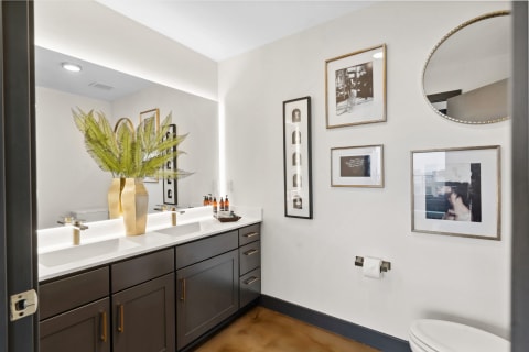 A stylish bathroom featuring a double vanity with greenery and framed art on the walls.