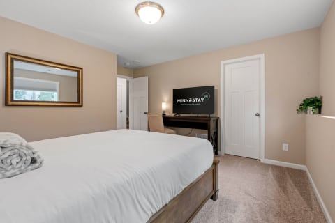 A stylish bedroom featuring a white bed, decorative mirror, television, and a comfortable chair.