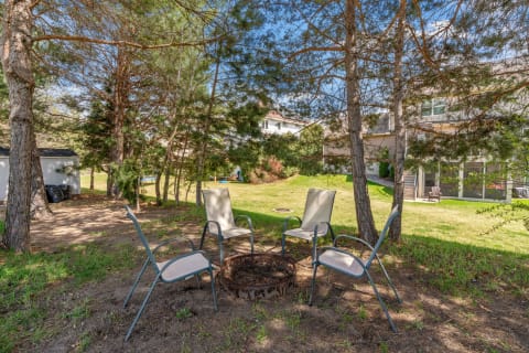 An outdoor fire pit with chairs in a green lawn, surrounded by trees and a house in the background.