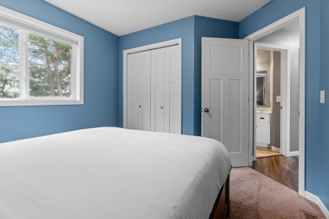 A cozy bedroom featuring blue walls and a white bedspread, with a large window and doors leading to a closet and bathroom.