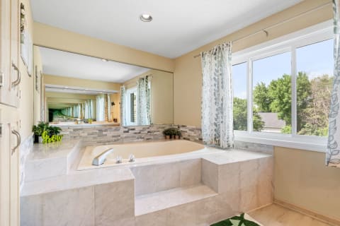 A bathroom with a marble bathtub, wooden cabinets, and botanical curtains, illuminated by natural light.