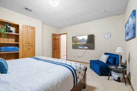 Cozy bedroom with a bed, blue lounge chair, and a mounted TV showing a landscape.