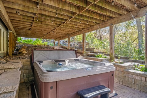 A backyard hot tub situated under a wooden structure, with surrounding greenery and stone features.
