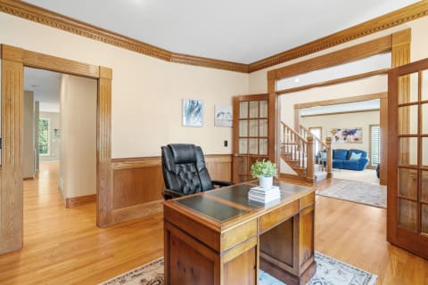 A home office with a wooden desk, black leather chair, and a view into a living room with a blue sofa.